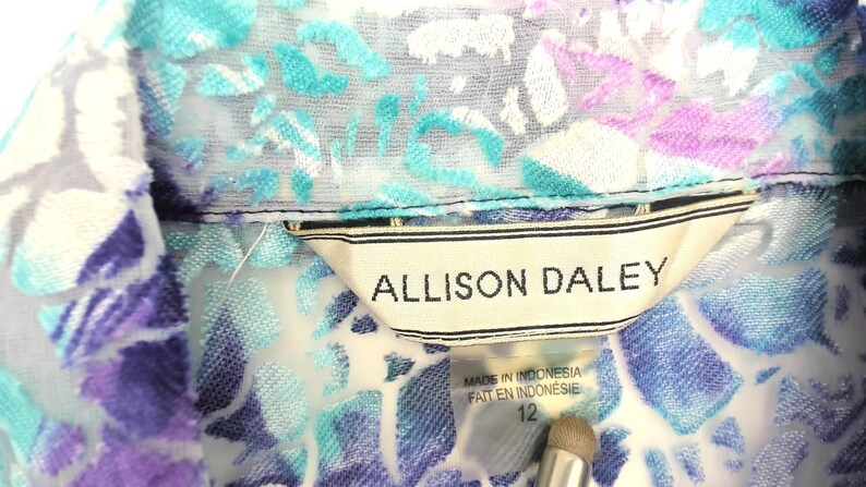 Op de afbeelding: Close-up van een blouse met bloemenpatroon en kraag. De blouse heeft een mix van blauwe, paarse en witte kleuren. Het label luidt "ALLISON DALEY" en "MADE IN INDONESIA". De maat is 12.