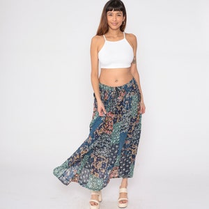 Op de afbeelding: Een vrouw draagt een wit crop top en een lange, blauw-groene patchwork rok met bloemenpatronen. Ze draagt witte sandalen.