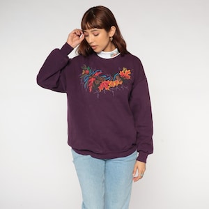 Op de afbeelding: Een donkerpaarse sweatshirt met een kleurrijk herfstbladontwerp. De afbeelding toont rode, oranje en gele bladeren met blauwe accenten. De sweatshirt heeft lange mouwen en een ronde hals.
