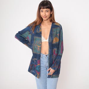 Könnte beinhalten: Ein blauer Vintage-Blazer mit einem farbenfrohen geometrischen Muster. Die offene Jacke hat einen V-Ausschnitt und lange Ärmel. Das Muster umfasst Farbtöne von Türkis, Lila und Gold. Der Blazer ist ein einzigartiges Vintage-Kleidungsstück.
