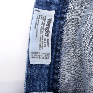 Puede incluir: Primer plano de unos vaqueros de mezclilla azules con una etiqueta Wrangler. La etiqueta indica la talla 34 x 33. Los vaqueros son 100% algod&oacute;n y est&aacute;n fabricados en M&eacute;xico con tejido estadounidense.