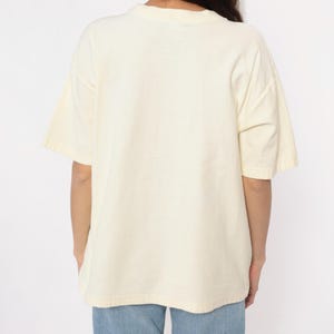 May include: Cream-colored short-sleeved t-shirt. The shirt has a crew neck and a relaxed fit. The fabric appears to be a soft, lightweight material. The model is wearing blue jeans.