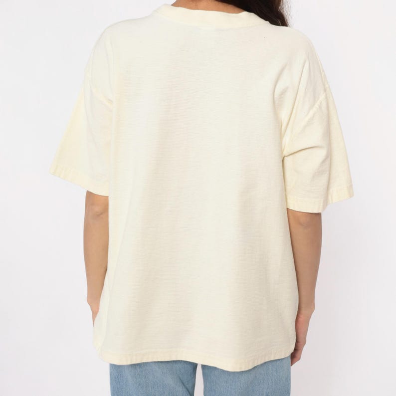 May include: Cream-colored short-sleeved t-shirt. The shirt has a crew neck and a relaxed fit. The fabric appears to be a soft, lightweight material. The model is wearing blue jeans.