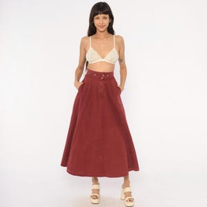 May include: A high-waisted, ankle-length skirt in a deep brick red color, featuring a button-front closure, a structured waistband, and side pockets. The skirt has a flared, A-line silhouette made from a sturdy fabric, paired with a cream lace bralette.