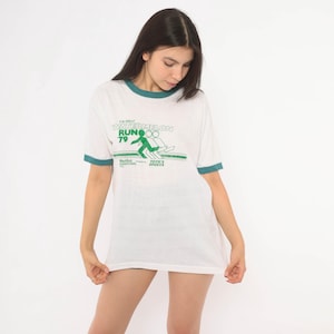 May include: A white ringer t-shirt with dark green trim on the collar and sleeves. The front features a green graphic with the text THE GREAT WATERMELON RUN '79, Nautilus Downtown Ltd, and Toth's Sports, depicting a stylized running figure.
