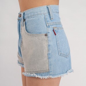 Puede incluir: Pantalones cortos de mezclilla azul claro con un bolsillo de parche gris contrastante en el lado derecho. Los pantalones cortos tienen un dobladillo deshilachado y una etiqueta roja en el bolsillo trasero.