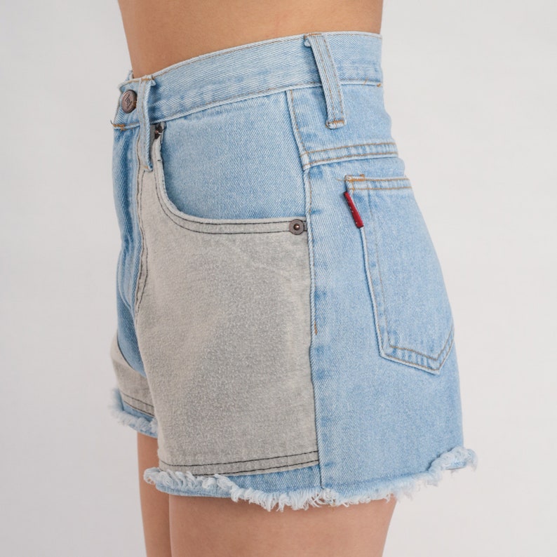 Puede incluir: Pantalones cortos de mezclilla azul claro con un bolsillo de parche gris contrastante en el lado derecho. Los pantalones cortos tienen un dobladillo deshilachado y una etiqueta roja en el bolsillo trasero.