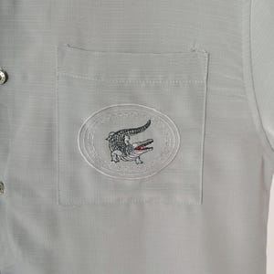 May include: A gray button-down shirt with a patch pocket featuring an embroidered alligator logo.