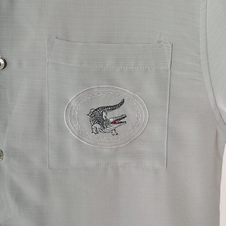 May include: A gray button-down shirt with a patch pocket featuring an embroidered alligator logo.