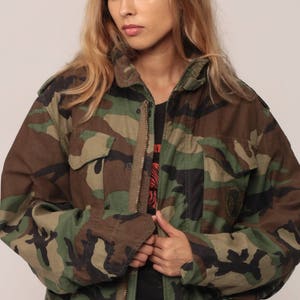 May include: A camouflage field jacket featuring a woodland pattern in shades of olive green, brown, and black. The jacket includes a high collar, a full-length front zipper, two chest pockets with buttoned flaps, and long sleeves with cuffs.