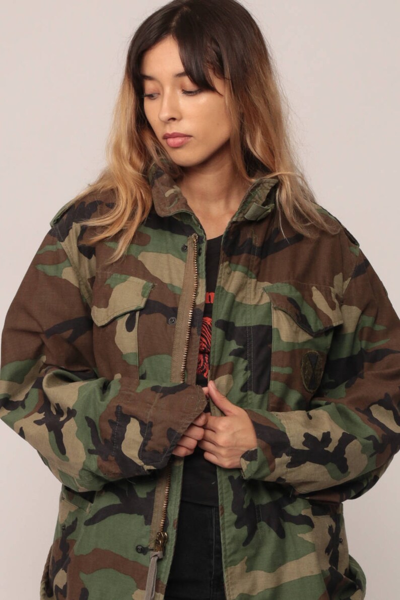 May include: A camouflage field jacket featuring a woodland pattern in shades of olive green, brown, and black. The jacket includes a high collar, a full-length front zipper, two chest pockets with buttoned flaps, and long sleeves with cuffs.