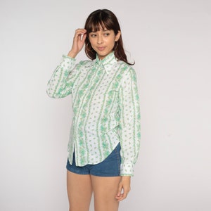 Puede incluir: Una camisa blanca de manga larga con un estampado floral verde y blanco. La camisa tiene cuello y cierre de botones en la parte delantera.