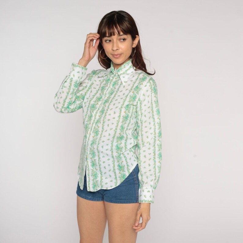 Puede incluir: Una camisa blanca de manga larga con un estampado floral verde y blanco. La camisa tiene cuello y cierre de botones en la parte delantera.