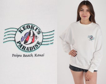Op de afbeelding: Witte trui met een blauw, groen en rood logo met de tekst "Keoki's Paradise" en "Poipu Beach, Kauai".