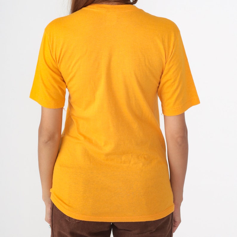 May include: Mustard yellow short-sleeve t-shirt. The back of the shirt is visible, showing the neckline and the short sleeves. The shirt is made of a soft, textured fabric. The model is wearing brown pants.