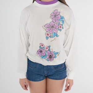 May include: White long-sleeved shirt with a floral design featuring purple, pink, and blue flowers. The flowers are embroidered on the shirt.