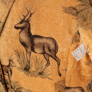 May include: Close-up of a tapestry featuring a detailed depiction of a deer with large antlers, set against a backdrop of tall grass and trees. The color palette includes shades of brown, tan, and green, creating a rustic aesthetic. The fabric has a woven texture.