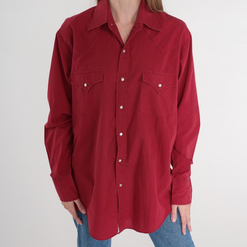 May include: A long-sleeved, red button-down shirt with a pointed collar and two chest pockets. The shirt has a Western style with pearl snaps.