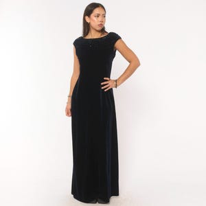 May include: A floor-length dark navy blue velvet gown featuring a boat neckline and short cap sleeves. The upper bodice and neckline are embellished with a dense pattern of small, dark, reflective beads that catch the light.