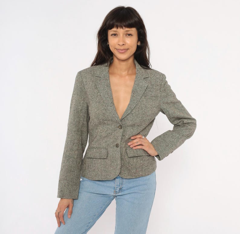 May include: A gray tweed blazer with a two-button closure, two front pockets, and a notched lapel. The blazer is paired with light blue jeans. The jacket has a textured appearance and a classic, tailored design.