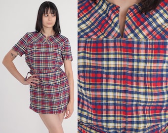 May include: A short-sleeved dress with a red, blue, and yellow plaid pattern. The dress has a V-neckline and a matching belt. The fabric appears to be lightweight, suitable for warmer weather. The dress is a vintage style.