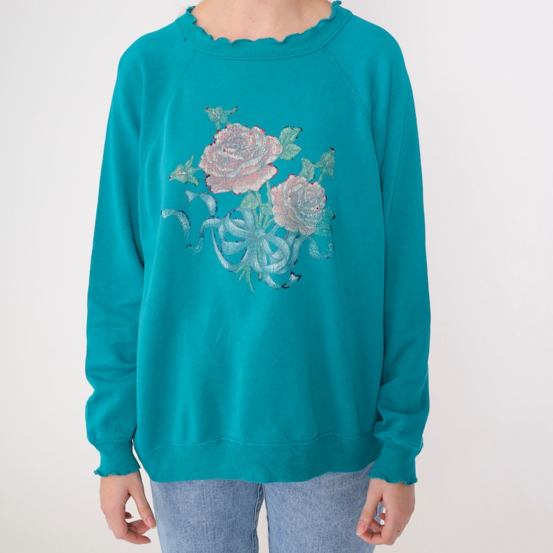 May include: A turquoise blue sweatshirt with a floral design featuring pink roses and green leaves. The sweatshirt has a ruffled neckline and long sleeves.