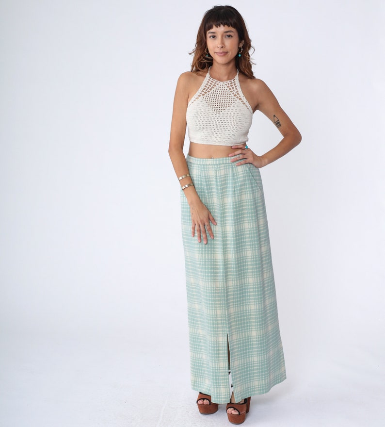 May include: A woman wearing a white crochet halter top and a long, light blue and white plaid skirt with a slit up the front. She is wearing brown heeled sandals.