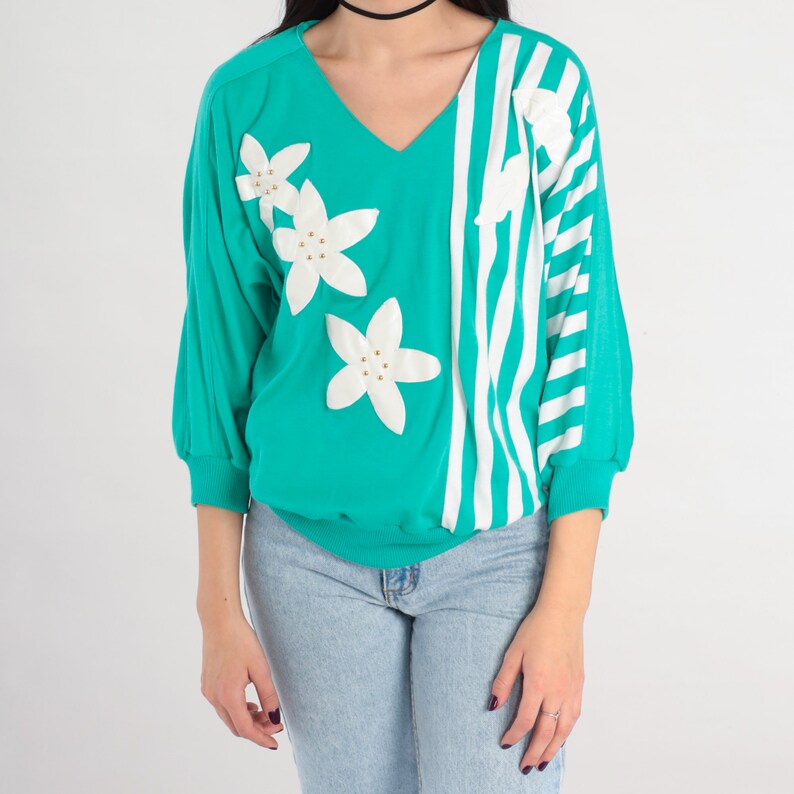 May include: A teal blue and white striped sweater with white flower embellishments. The sweater has a v-neck and three-quarter sleeves.