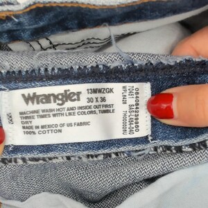 以下が含まれることがあります： 青いデニムのWranglerジーンズのクローズアップ。ラベルにはサイズ30 x 36インチと記載されています。ジーンズは綿100％で、メキシコ製です。ラベルには「Wrangler」と記載されています。