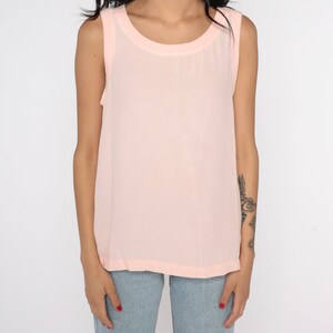 May include: A sleeveless, pale pink tank top with a rounded neckline. The top is made of a lightweight, semi-sheer fabric. The model is wearing blue jeans.