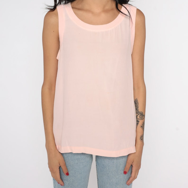 May include: A sleeveless, pale pink tank top with a rounded neckline. The top is made of a lightweight, semi-sheer fabric. The model is wearing blue jeans.