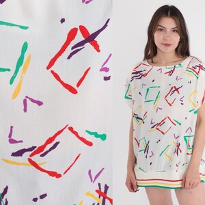 Puede incluir: Top blanco de manga corta con un colorido estampado geométrico. El diseño presenta formas rojas, verdes, amarillas y moradas. El top tiene un cuello redondo y un borde a rayas en el dobladillo.
