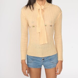 May include: A pale yellow ribbed knit sweater with a tie at the neck and a floral embroidered design. The sweater has long sleeves and a fitted silhouette.