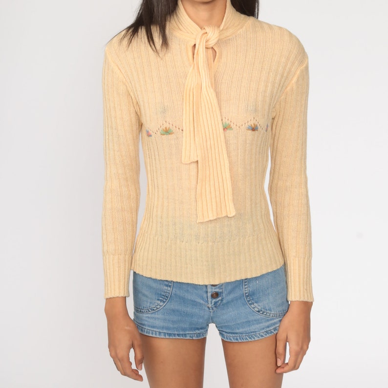 May include: A pale yellow ribbed knit sweater with a tie at the neck and a floral embroidered design. The sweater has long sleeves and a fitted silhouette.