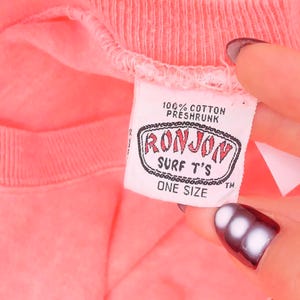 K&ouml;nnte beinhalten: Ein leuchtend korallenrotes T-Shirt mit einem wei&szlig;en Innenetikett am Kragen. Das Etikett enth&auml;lt den Text 100% COTTON PRESHRUNK, RONJON SURF T'S und ONE SIZE. Der Stoff hat einen gerippten Kragen und eine leicht texturierte Optik am gesamten Kleidungsst&uuml;ck.