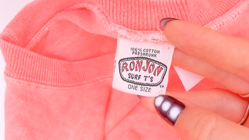 K&ouml;nnte beinhalten: Ein leuchtend korallenrotes T-Shirt mit einem wei&szlig;en Innenetikett am Kragen. Das Etikett enth&auml;lt den Text 100% COTTON PRESHRUNK, RONJON SURF T'S und ONE SIZE. Der Stoff hat einen gerippten Kragen und eine leicht texturierte Optik am gesamten Kleidungsst&uuml;ck.