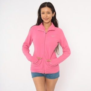 May include: A bright pink long-sleeve fleece jacket featuring a full-length front zipper, a high collar, and two front pockets. The garment displays a small, silver-toned Nike swoosh logo on the upper left chest area, providing a subtle contrast.