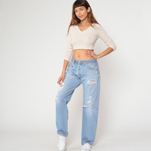 May include: Light blue denim jeans with distressed details, paired with a cream-colored, long-sleeved, v-neck crop top. The jeans have a button fly and multiple rips. White sneakers complete the casual outfit.