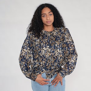 May include: A long-sleeved blouse with a blue, tan, and black abstract pattern. The blouse has a round neckline, gathered cuffs, and a cinched waist. The model is wearing light blue jeans.
