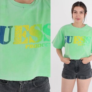 May include: A light green cropped t-shirt with a multi-coloured "GUESS PRODUCT" logo printed on the front.