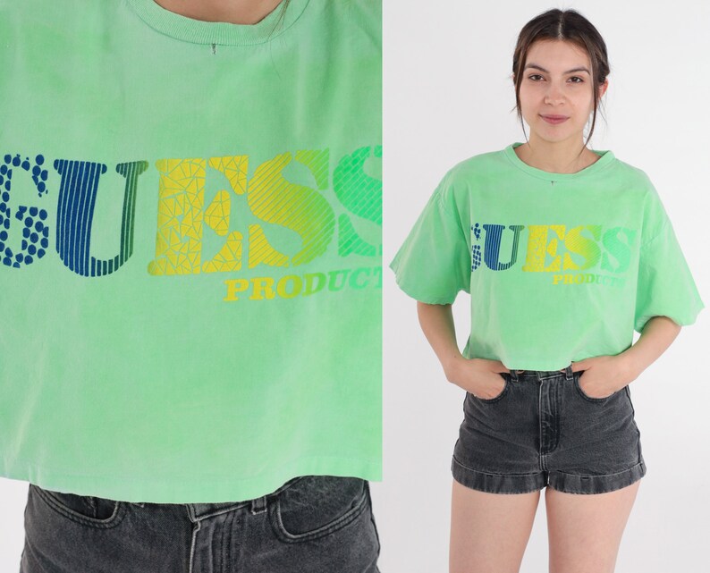 May include: A light green cropped t-shirt with a multi-coloured "GUESS PRODUCT" logo printed on the front.