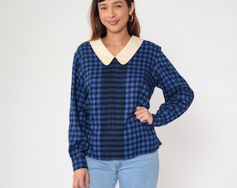 Può includere: Una camicetta a maniche lunghe a quadri blu e neri con colletto Peter Pan color crema. La camicetta è abbinata a jeans azzurri. La donna indossa orecchini a cerchio dorati.