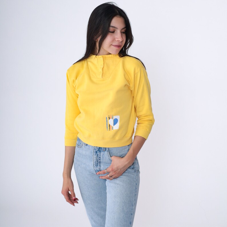 May include: A yellow long-sleeved shirt with a button-up collar and a small blue heart design on the left side.
