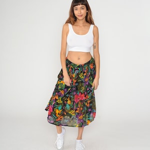 May include: A woman wearing a white crop top and a floral print skirt. The skirt features a black background with colorful flowers in shades of pink, orange, yellow, and blue. The outfit is paired with white sneakers.
