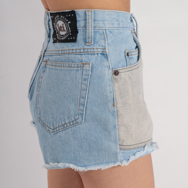 Puede incluir: Pantalones cortos de mezclilla azul claro con dobladillo deshilachado y un bolsillo de parche gris contrastante en la parte posterior. Los pantalones cortos tienen una etiqueta negra con el texto "Long Wear Quality Product" y un logotipo.