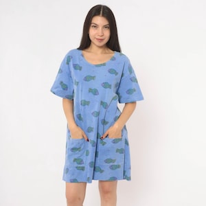 May include: A light blue short-sleeved dress featuring a repeating pattern of small green and purple fish. The garment has a round neckline and two large square patch pockets on the front, providing a relaxed fit that reaches above the knees.