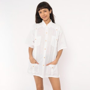 May include: A white short-sleeved button-down shirt featuring a classic collar, two chest pockets, and two lower front pockets. The shirt is constructed with vertical pleats running down the front and is fastened with small, light-colored buttons.