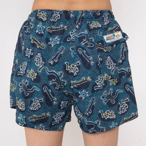 May include: Blue swim trunks with a pattern of green turtles, alligators, and lizards on a blue background with white dots. The trunks have a small pocket on the back with a label that says "Beach Bum" in yellow and red letters.