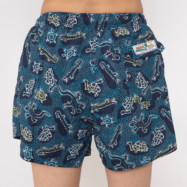 May include: Blue swim trunks with a pattern of green turtles, alligators, and lizards on a blue background with white dots. The trunks have a small pocket on the back with a label that says "Beach Bum" in yellow and red letters.