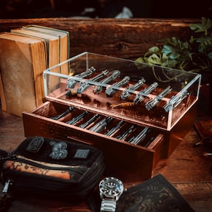 May include: A wooden box with a clear acrylic top and a drawer. The box is filled with a collection of small, silver-colored pocket knives. The box is sitting on a wooden table.